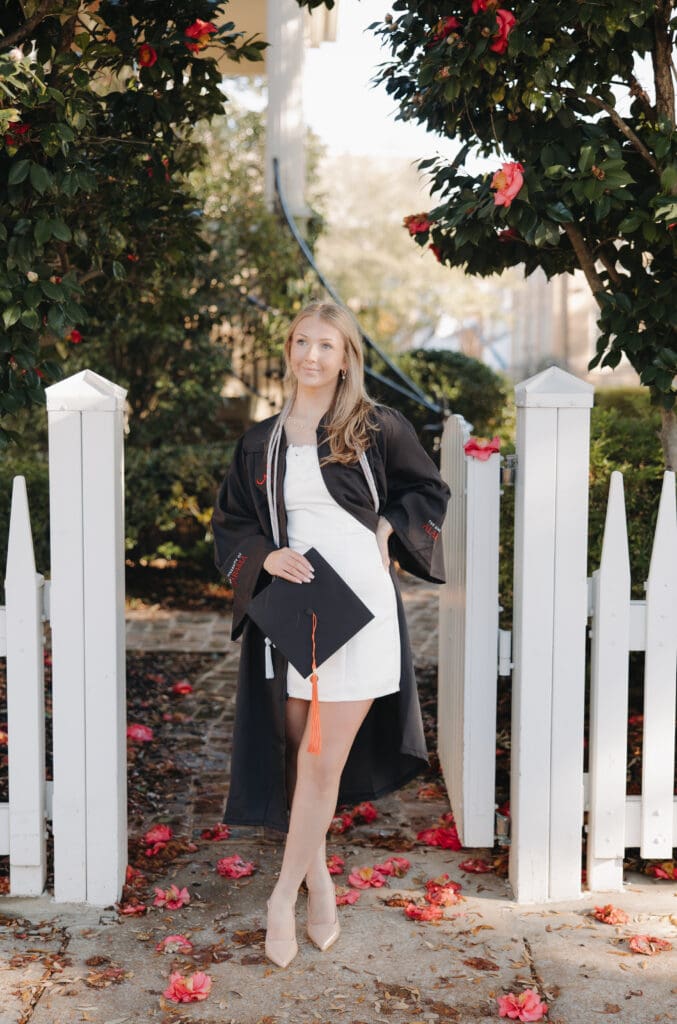 University of Alabama graduate in cap and gown standing between camellia trees in Tuscaloosa Alabama during senior photos